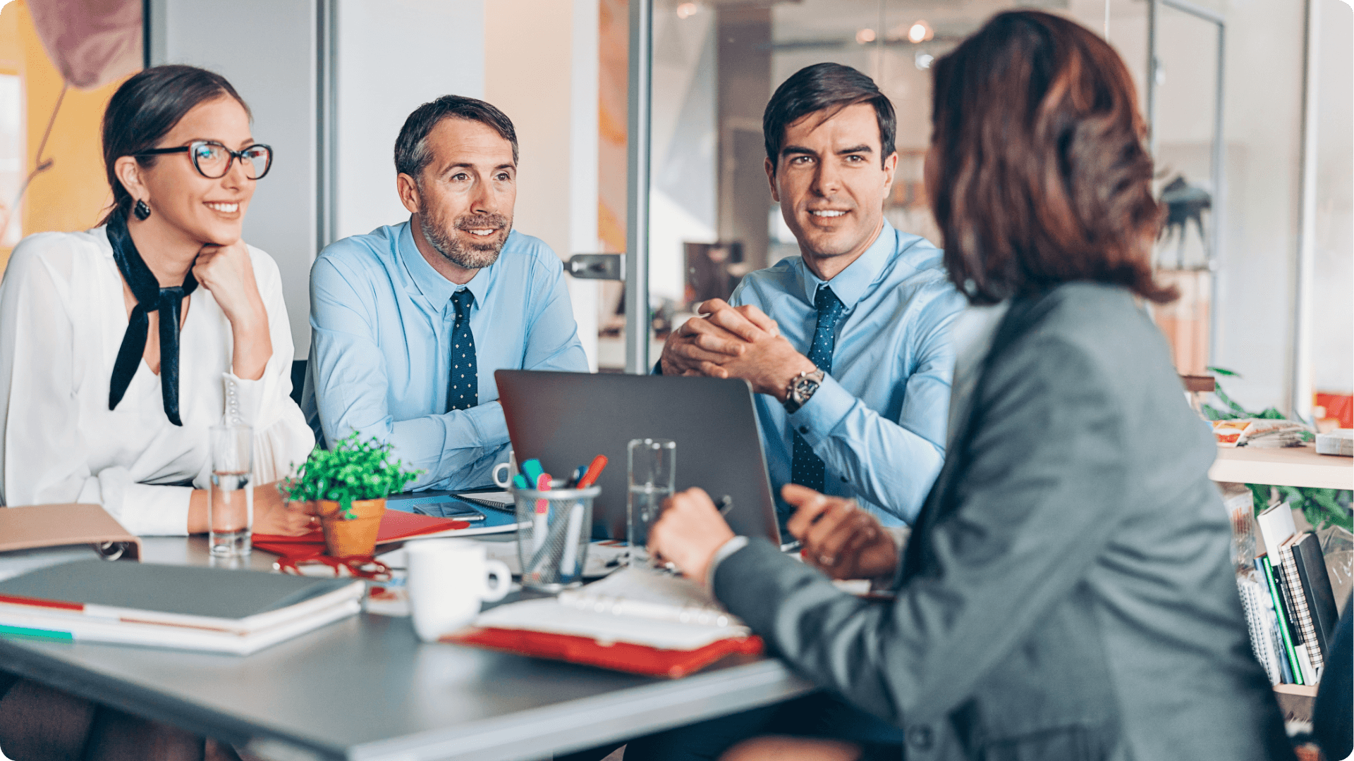 A group of It workers having a conversation around a table with a computer and multiple notebooks on the table in an office. (1)